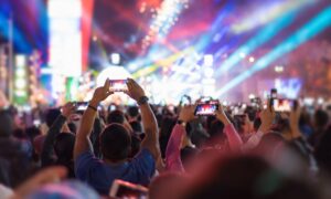 Imagem de várias pessoas com os braços levantados segurando o smartphone tirando foto de um palco grande com luzes ao fundo.